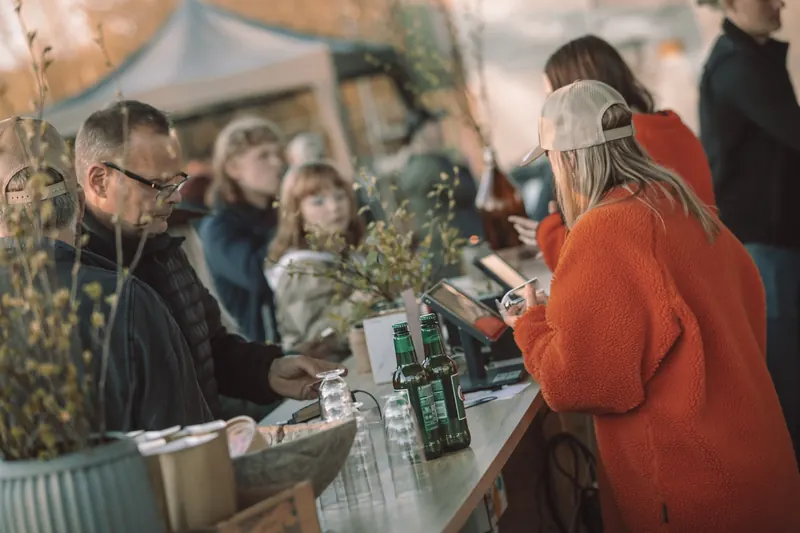 Besökare och personal vid den långa bardisken med ölflaskor och björkris i kvällssolen på Backwoods 2026