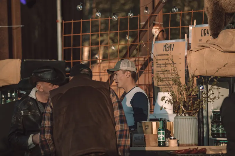 Besökare i cowboyhatt och flanellskjorta vid baren, med menyn uppklämd på armeringsnät i kvällssolen, på Backwoods 2026