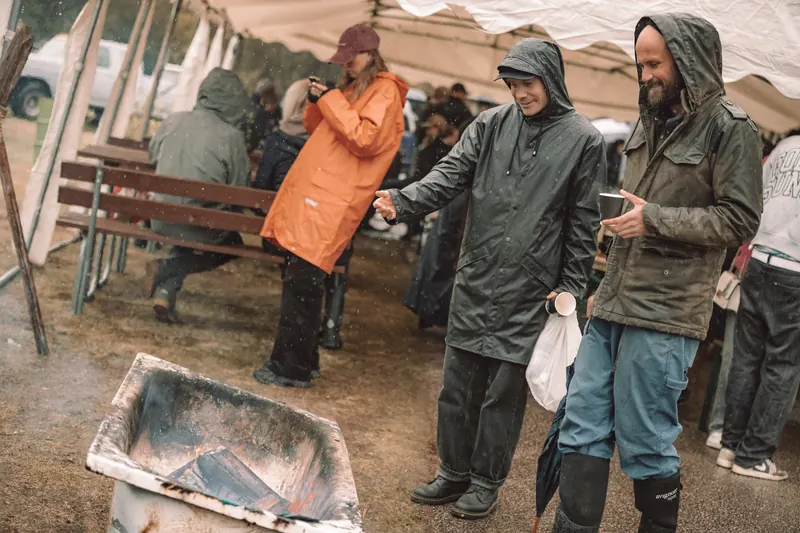 Två besökare i regnjackor skrattar och värmer sig vid en eld i ett gammalt badkar under ett tält på Backwoods 2026