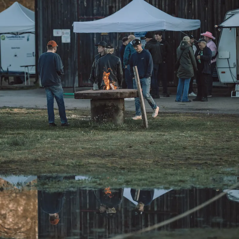Besökare samlas kring en eld i en gammal kvarnsten, speglad i en vattenpöl, framför sågverket på Backwoods 2026