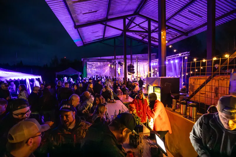 Besökare vid långborden och baren under det lila upplysta sågverkstaket på natten, med scenen i bakgrunden, på Backwoods 2026