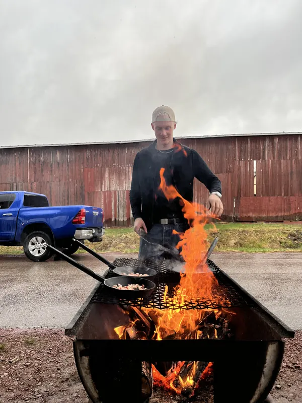 Matlagning i stekpannor över öppen eld i en eldstad, med en röd lada i bakgrunden, på Backwoods 2026