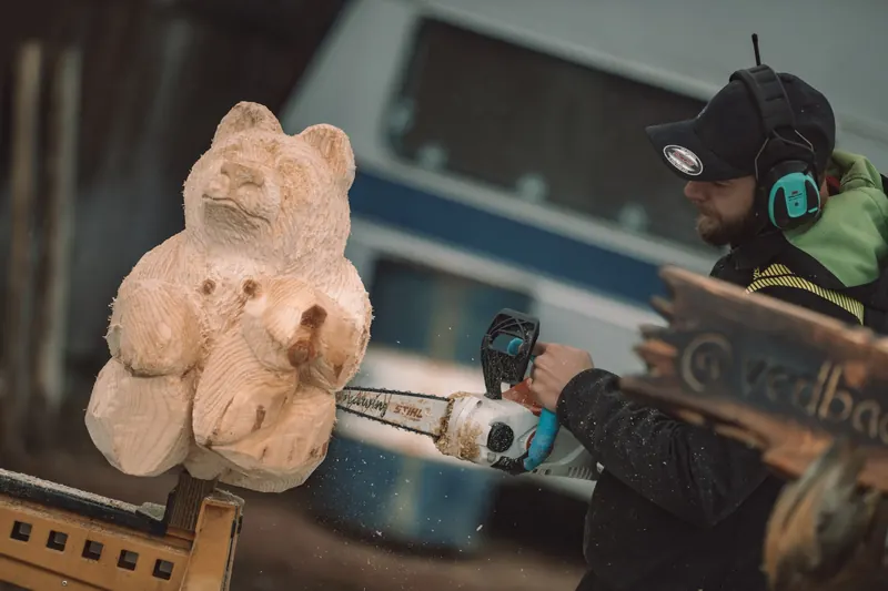 Närbild på en motorsågskonstnär med hörselskydd som sågar fram en björn ur en trästock på Backwoods 2026