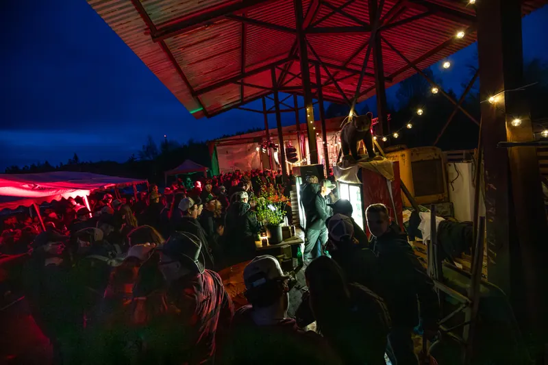 Tät publik under det rött upplysta sågverkstaket på natten, med den uppstoppade björnen ovanpå baren, på Backwoods 2026