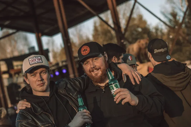 Två festivalbesökare i truckerkepsar skålar med ölflaskor under sågverkstaket, med den uppstoppade björnen i bakgrunden, på Backwoods 2026