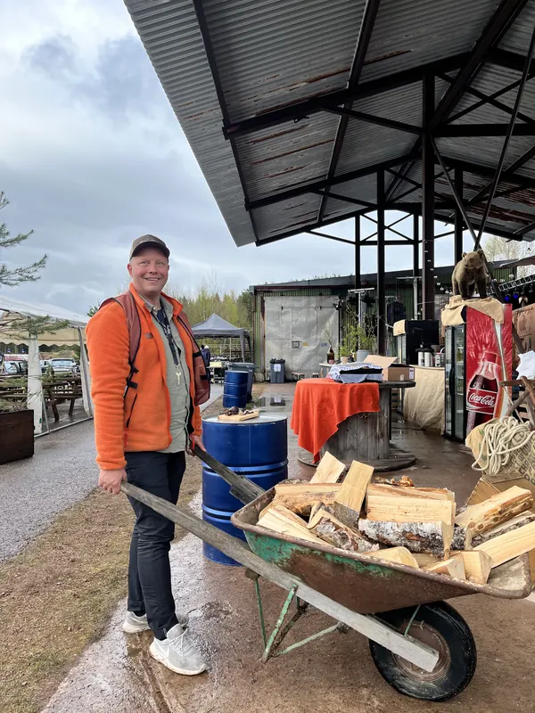 En man i orange jacka med en skottkärra full av ved under sågverkstaket på Backwoods 2026, med baren och den uppstoppade björnen bakom