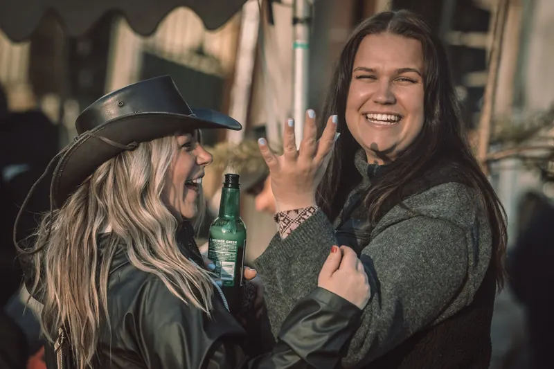 Två festivalbesökare skrattar tillsammans på Backwoods 2026, den ena i cowboyhatt med en ölflaska i handen