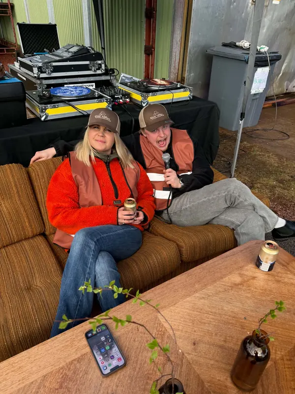Två personer i orange västar och kepsar sitter i en brun 70-talssoffa framför DJ-båset på Backwoods 2026, den ena med en mikrofon