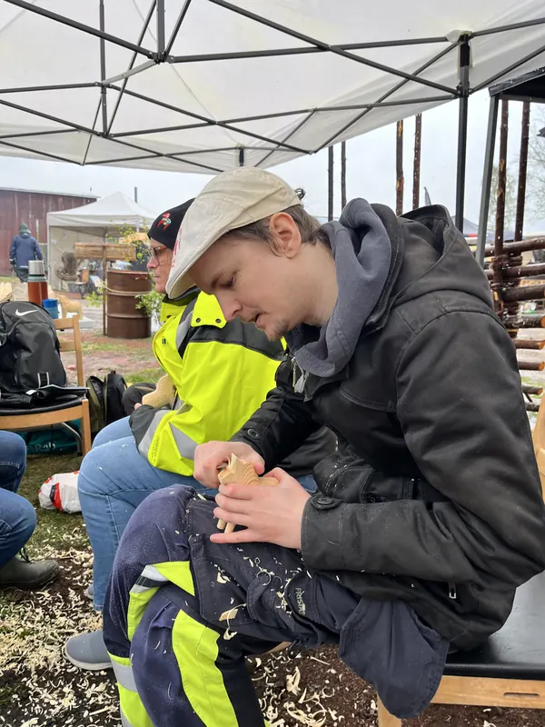 En ung man täljer i trä med spånen i knät under ett tält på Backwoods 2026