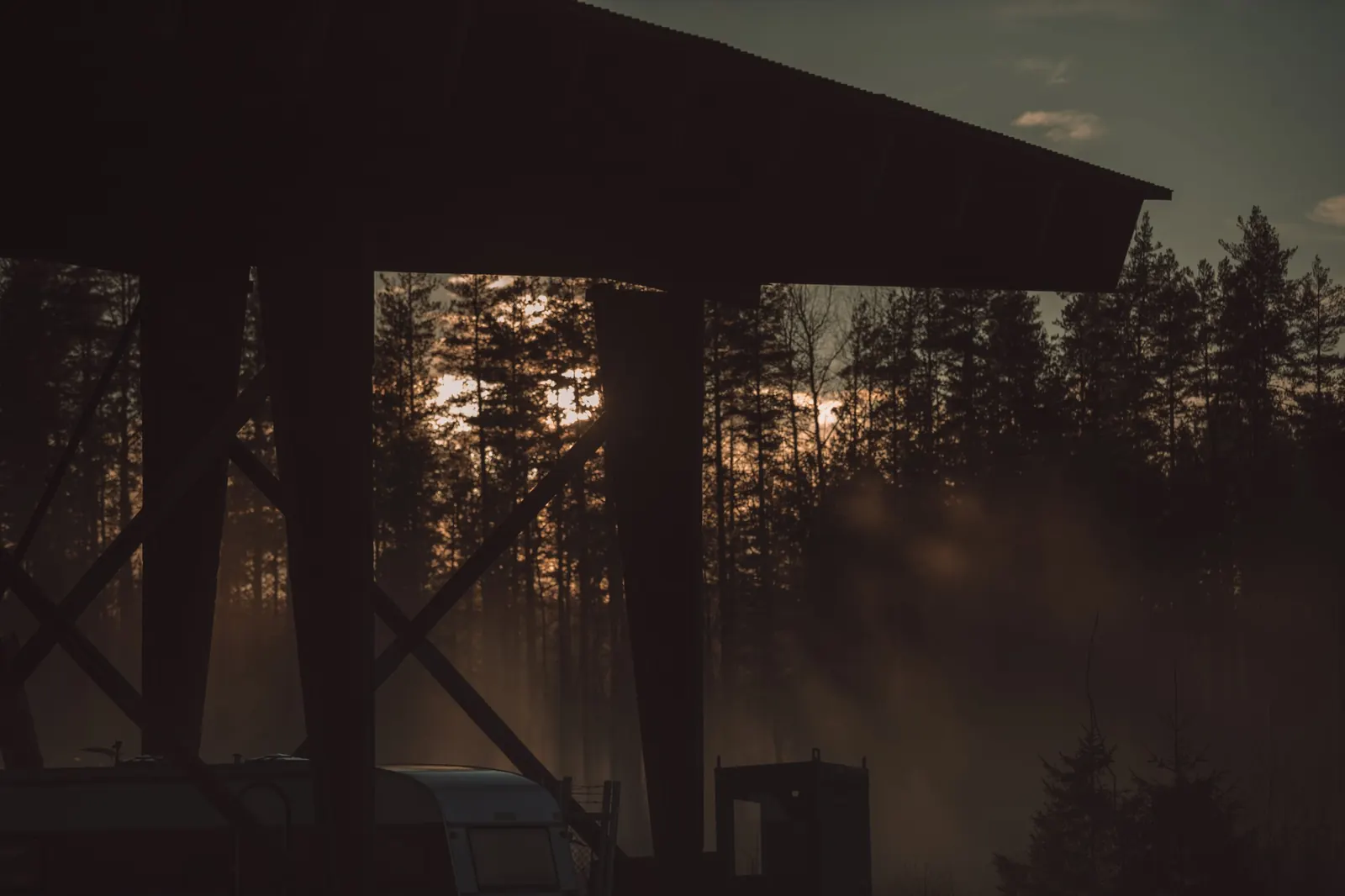Sågverkstaket på Rotsågen i siluett mot kvällssolen, med dimma mellan tallarna